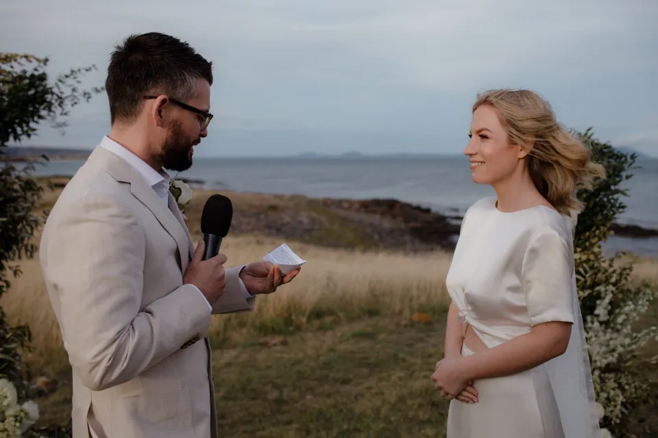 Jack speaking into a microphone as Jemmah listens to his vows, at Thalia Haven on the East Coast of Tasmania, Australia, for an Elopement Collective elopement. Photograph by Joey Willis.