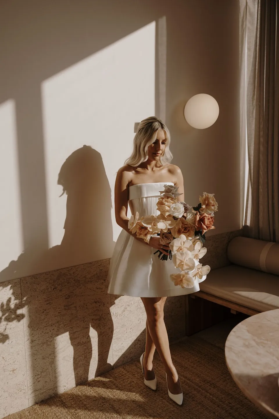 Danielle in her short white dress holding her bouquet by Mrs Gibbons Florals beside a sunlit suite window, at The Calile Hotel in Fortitude Valley, Brisbane, for an Elopement Collective elopement. Photo by George Bowden.