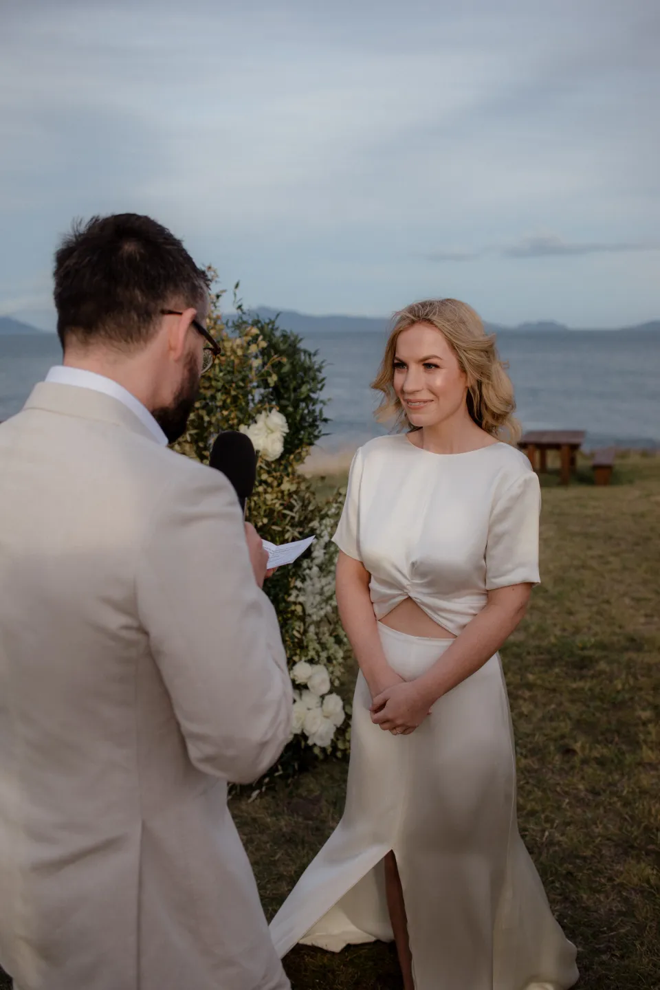 Jemmah smiling as Jack reads his vows beside the flowers by The Romantics, at Thalia Haven on the East Coast of Tasmania, Australia, for an Elopement Collective elopement. Photograph by Joey Willis.