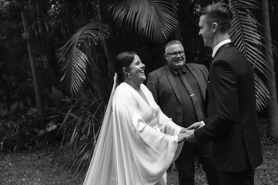 Black-and-white photograph of Laura smiling at Matthew during their ceremony with Josh Withers. At Fig Tree Villa in Ewingsdale, Byron Bay, for an Elopement Collective elopement. Photograph by George Bowden.