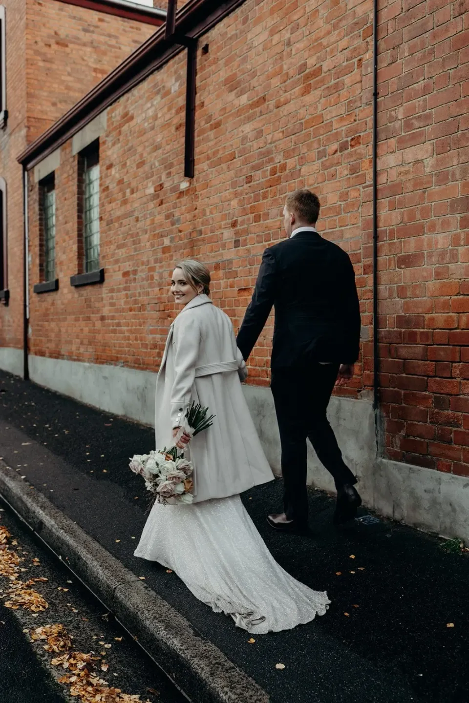 A couple walk hand in hand beside a red brick wall, the bride's train trailing behind.