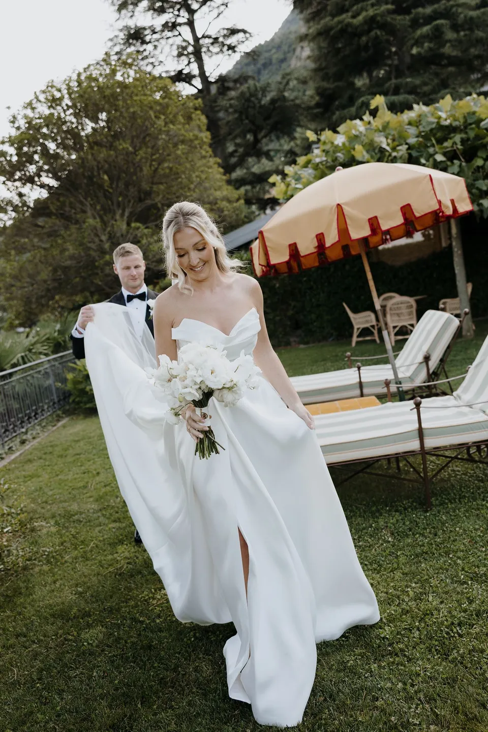 Zoe carrying her white bouquet by Garden of Love across the lawn, her veil trailing behind her beside a striped garden umbrella, at Passalacqua in Moltrasio, Lake Como, Italy, for an Elopement Collective elopement. Photo by George Bowden.