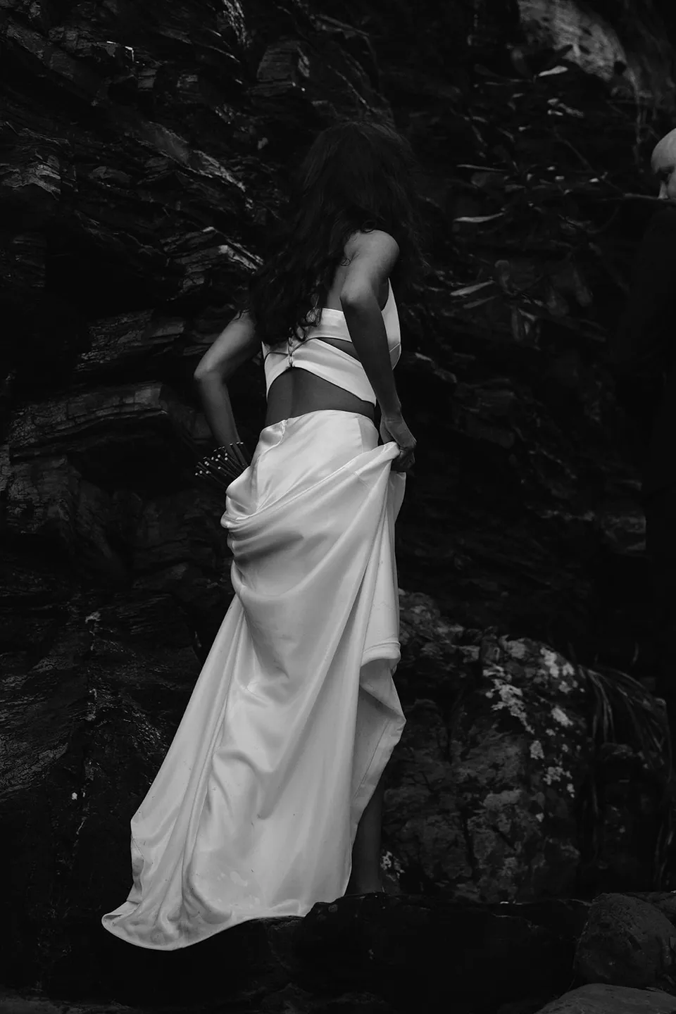 Black-and-white portrait of Rachel gathering her dress beside dark coastal rocks. At Whites Beach, Broken Head, near Byron Bay, for an Elopement Collective elopement. Photograph by Jason Lucas.