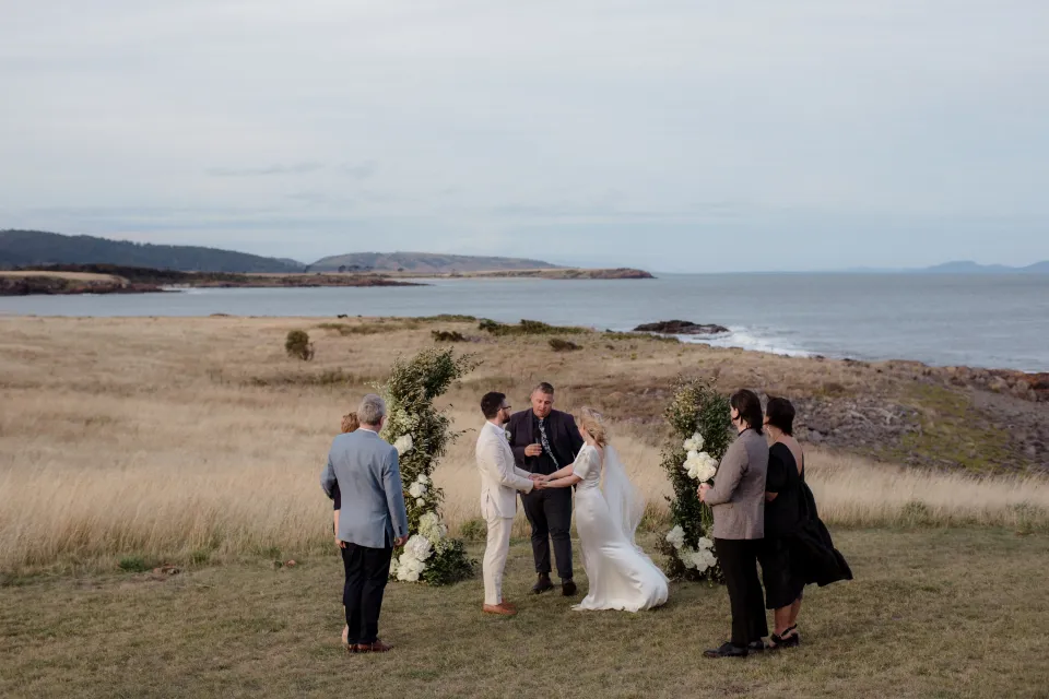 A wide view of the couple, Josh Withers and loved ones during the ceremony, at Thalia Haven on the East Coast of Tasmania, Australia, for an Elopement Collective elopement. Photograph by Joey Willis.