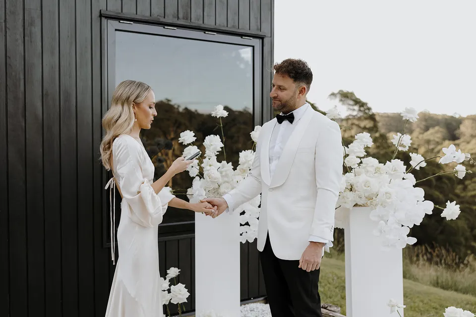 Gabrielle reading her vows while holding Greg’s hand beside the cabin’s dark timber wall, at Roundhill Retreat in Obi Obi, Sunshine Coast Hinterland, Queensland, for an Elopement Collective elopement. Photo by George Bowden.