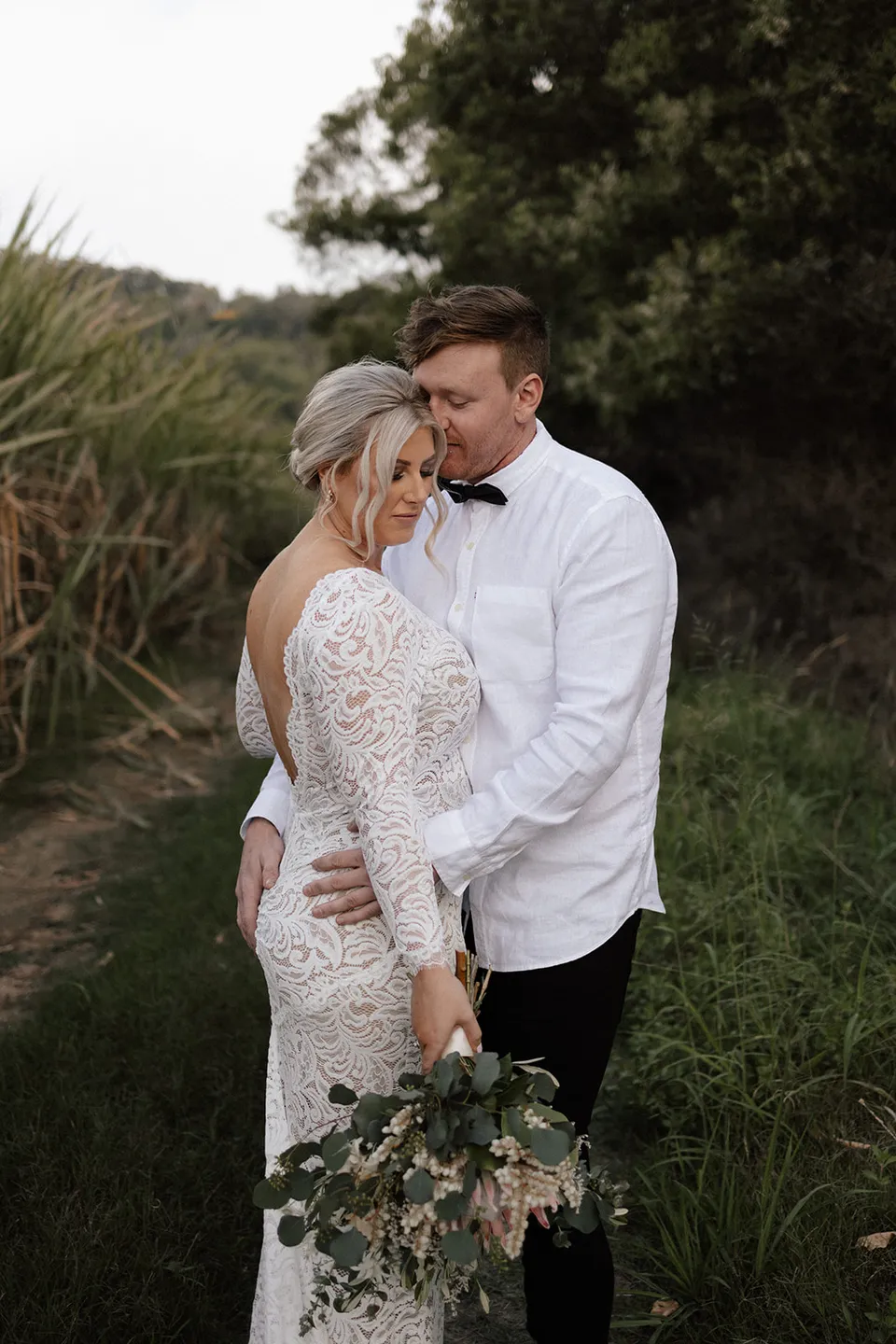 Matt embracing Laura beside tall grasses as she holds her bouquet by Fifth Avenue Florist, in the Tweed Valley, NSW, for an Elopement Collective elopement. Photo by Bulb Creative.