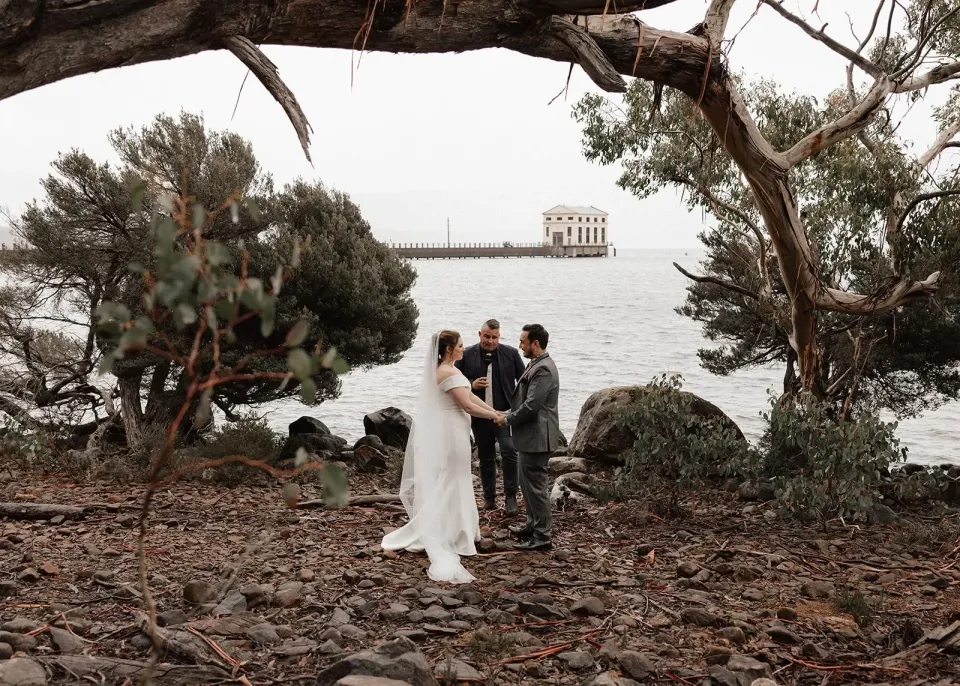 Sarah and Jono holding hands during their lakeside ceremony with celebrant Josh Withers beneath spreading trees, at Pumphouse Point, Lake St Clair, Tasmania, for an Elopement Collective elopement. Photo by Bulb Creative.