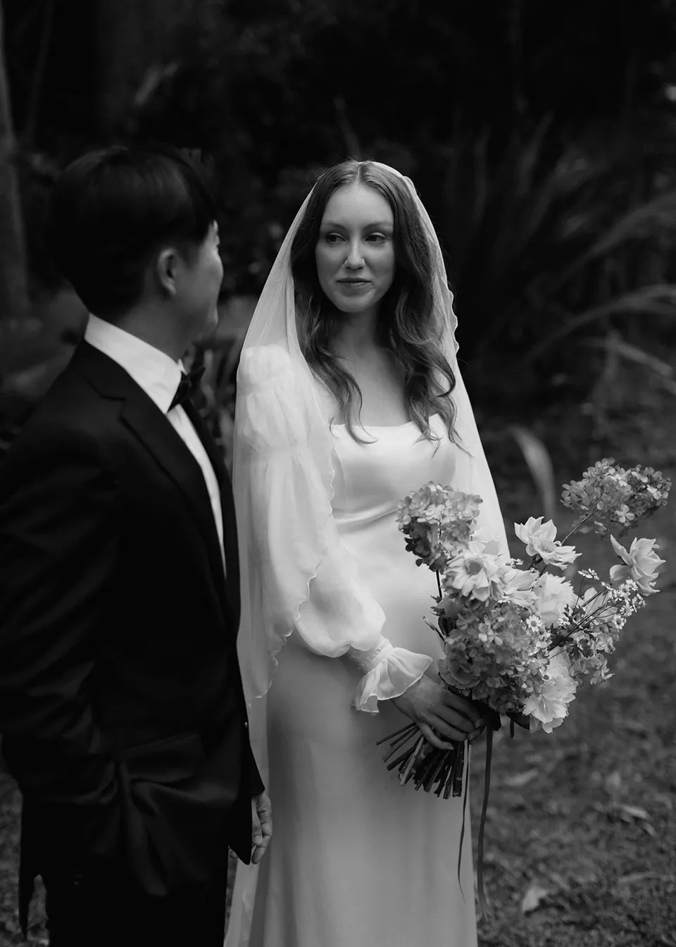 Black-and-white portrait of Elyse looking towards Jonathan with her bouquet, with flowers by The Perfect Party Co. At Fig Tree Villa in Ewingsdale, Byron Bay, for an Elopement Collective elopement. Photograph by Bulb Creative.