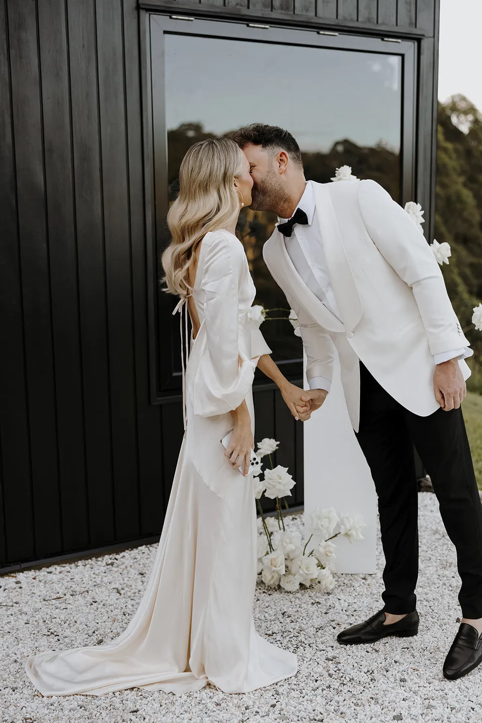 Gabrielle and Greg kissing beside the white ceremony flowers by Mrs Gibbons and cabin window, at Roundhill Retreat in Obi Obi, Sunshine Coast Hinterland, Queensland, for an Elopement Collective elopement. Photo by George Bowden.