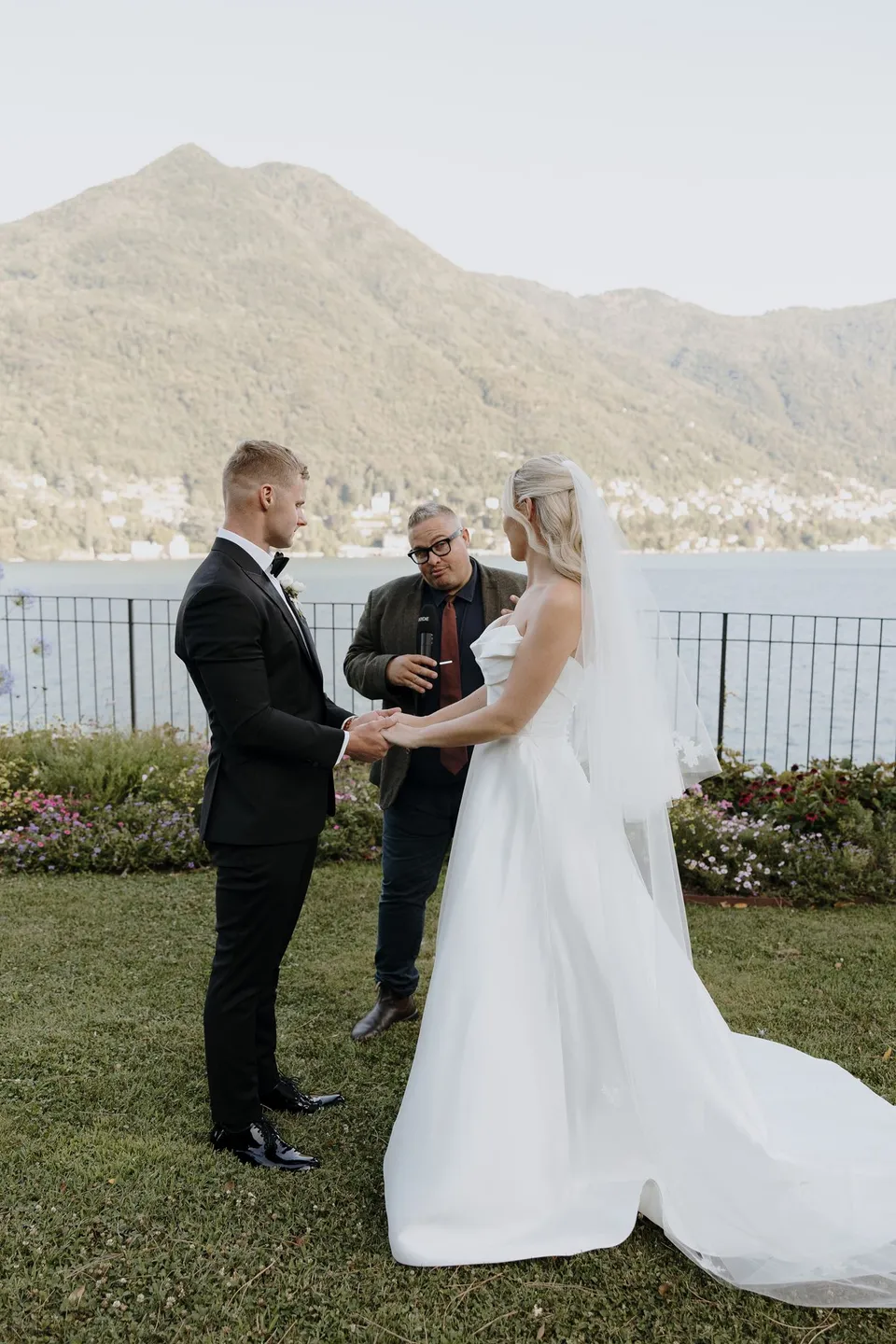 Celebrant Josh Withers leading Zoe and Charlton’s ceremony on a lawn above the lake, with the mountain slopes behind them, at Passalacqua in Moltrasio, Lake Como, Italy, for an Elopement Collective elopement. Photo by George Bowden.