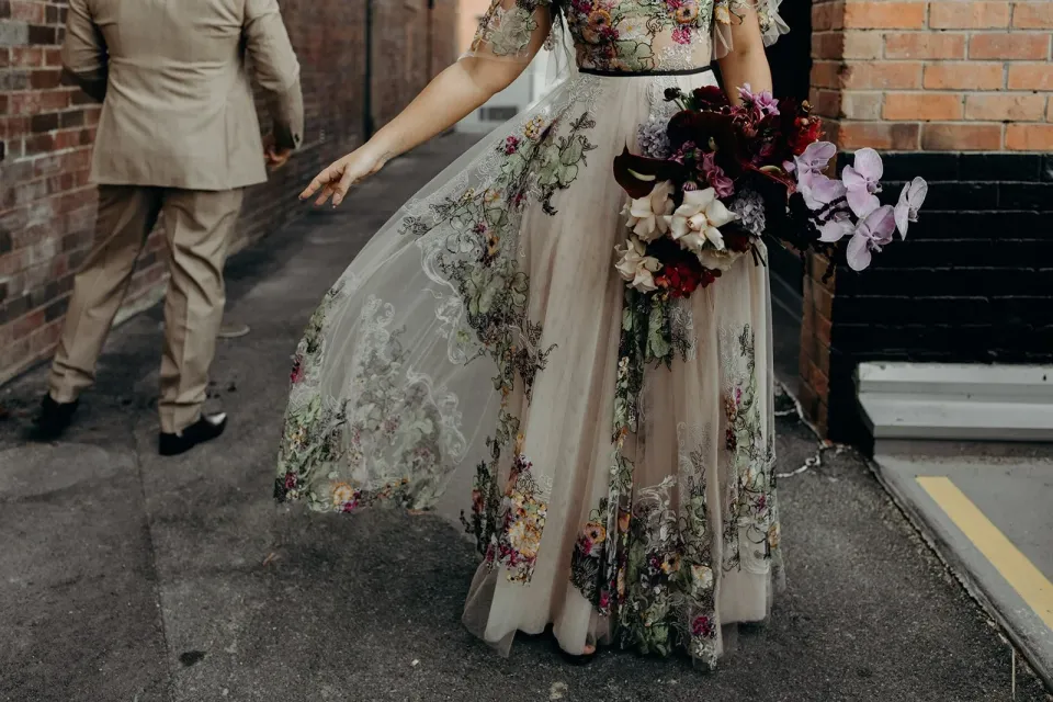 Victoria lifting the sheer floral layer of her skirt while holding her bouquet by Mrs Gibbons Flowers, with Daniel walking ahead, in Fortitude Valley, Brisbane, for an Elopement Collective elopement. Photo by Tessa Shannon.
