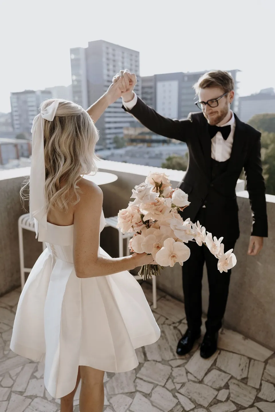 Adam lifting Danielle’s hand as she turns beside him on the terrace, carrying her bouquet by Mrs Gibbons Florals, at The Calile Hotel in Fortitude Valley, Brisbane, for an Elopement Collective elopement. Photo by George Bowden.