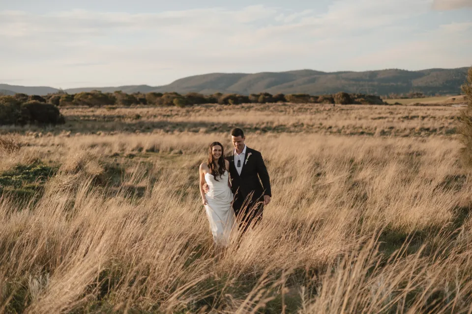 Dani and Anthony walking together through tall golden grass, with low coastal hills behind them, at Thalia Haven on Tasmania’s east coast, for an Elopement Collective elopement. Photograph by Bulb Creative.