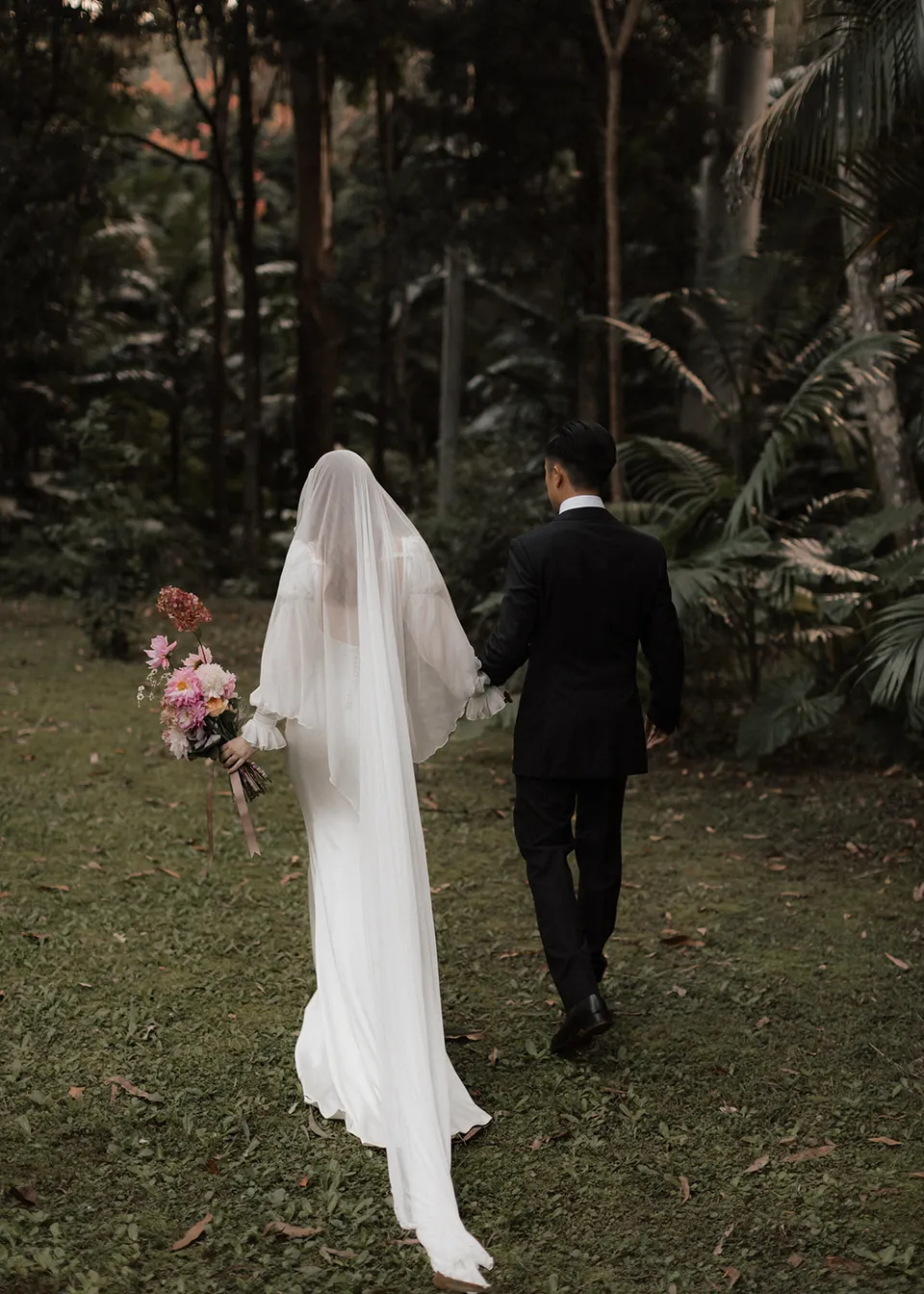 Rear view of the couple walking hand in hand towards the trees. At Fig Tree Villa in Ewingsdale, Byron Bay, for an Elopement Collective elopement. Photograph by Bulb Creative.