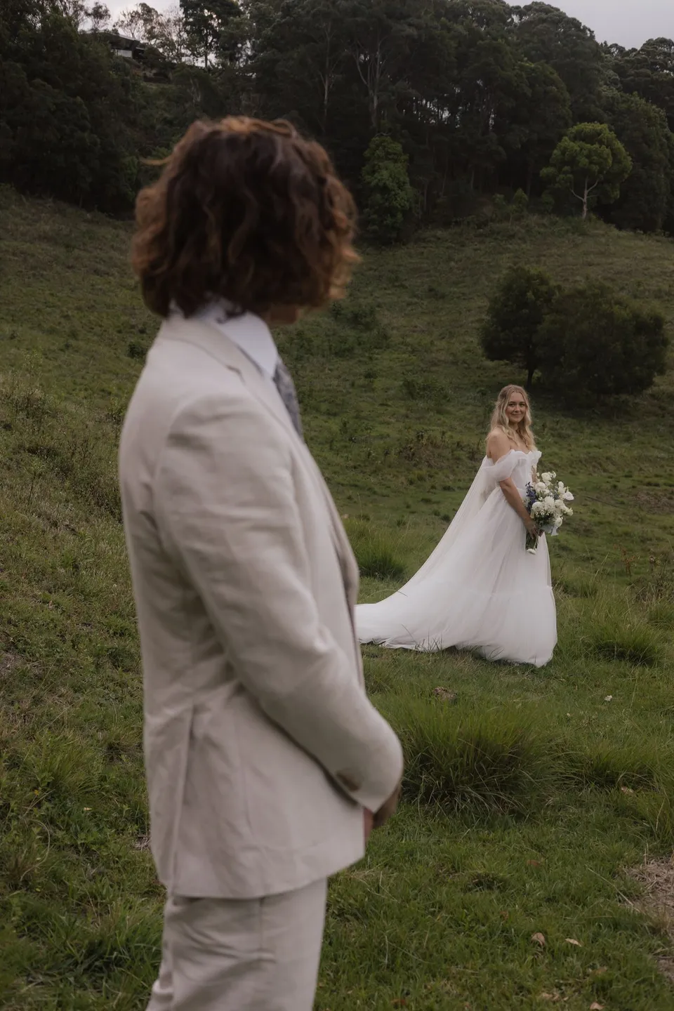 James looking back towards Bridget as she stands on the green hillside. At Cudgera Creek in northern New South Wales, for an Elopement Collective elopement. Photograph by Bulb Creative.