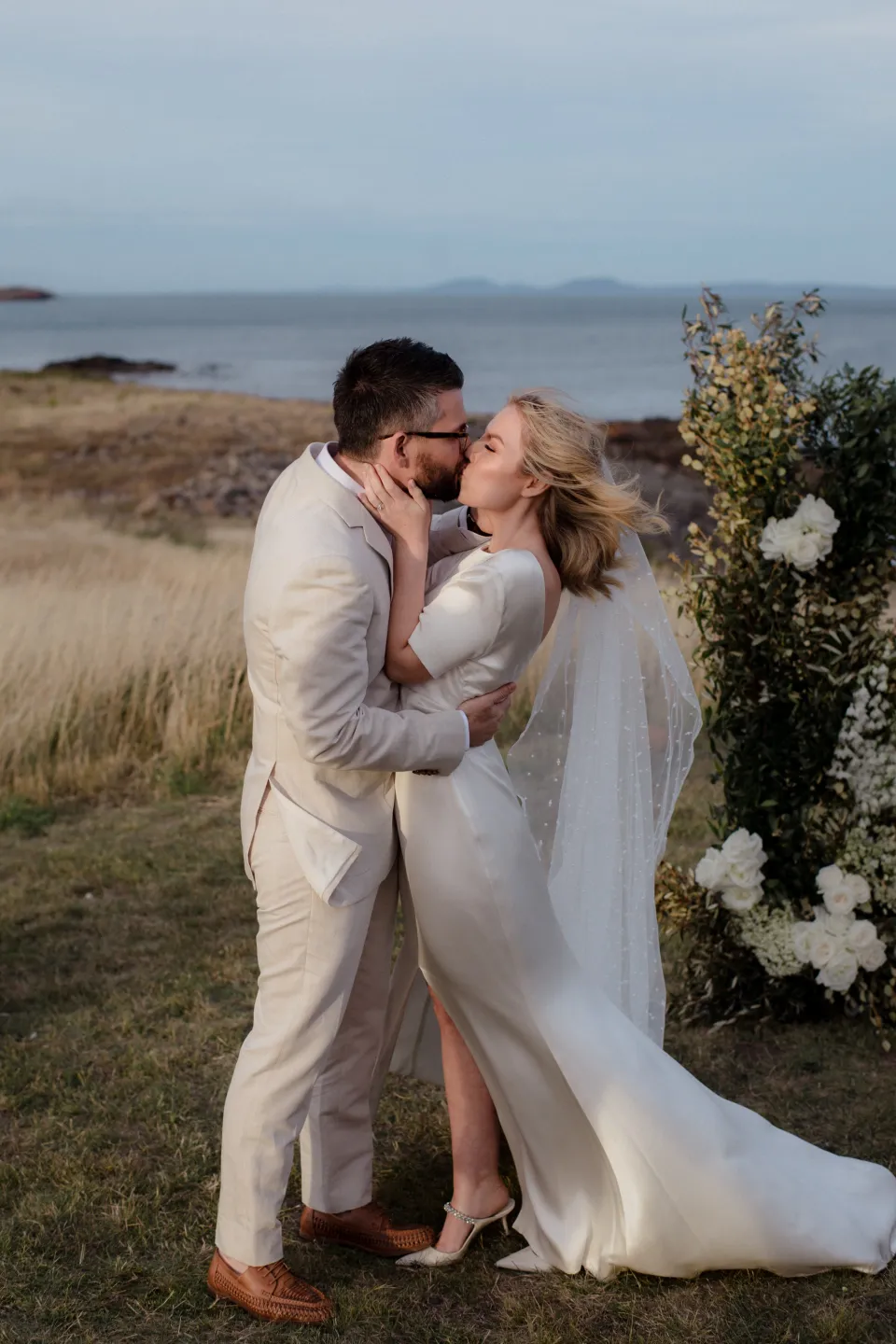 Jemmah and Jack kissing between the ceremony flowers by The Romantics, at Thalia Haven on the East Coast of Tasmania, Australia, for an Elopement Collective elopement. Photograph by Joey Willis.