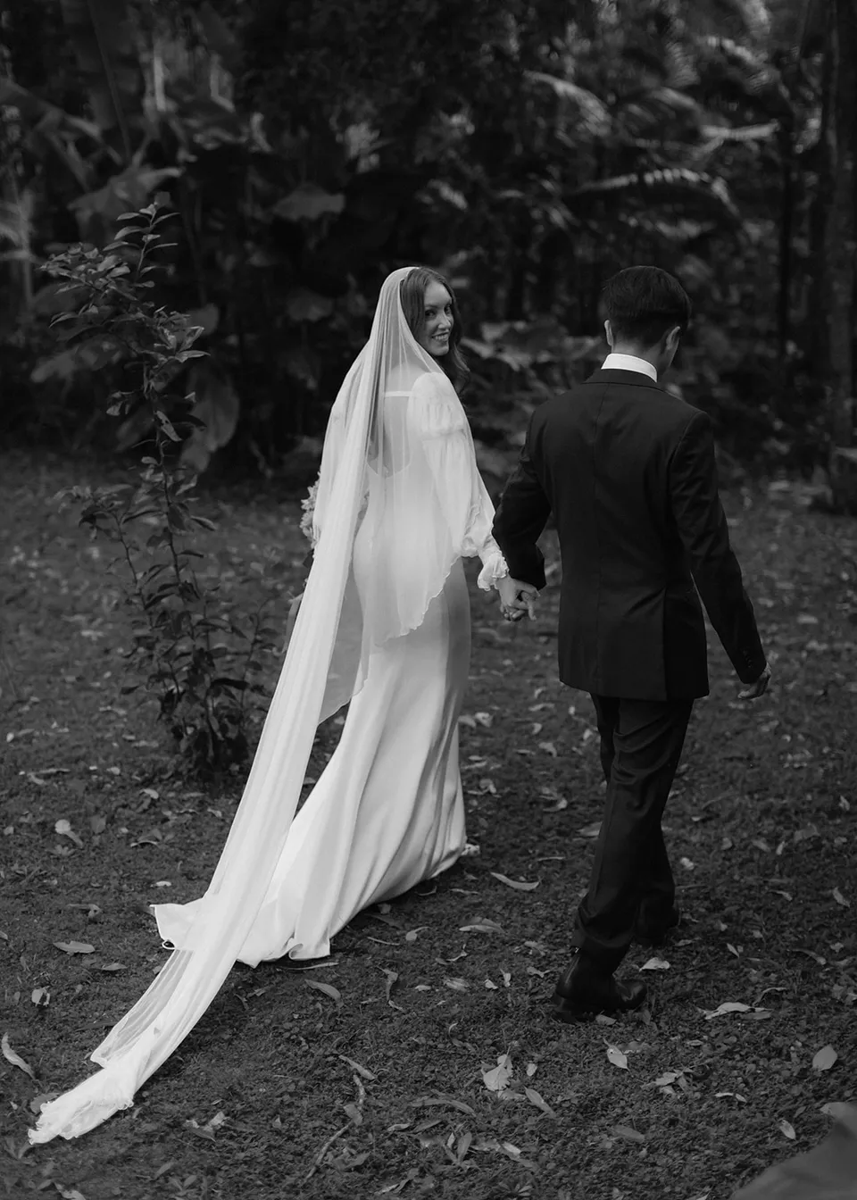 Black-and-white photograph of Elyse looking back while holding Jonathan's hand. At Fig Tree Villa in Ewingsdale, Byron Bay, for an Elopement Collective elopement. Photograph by Bulb Creative.