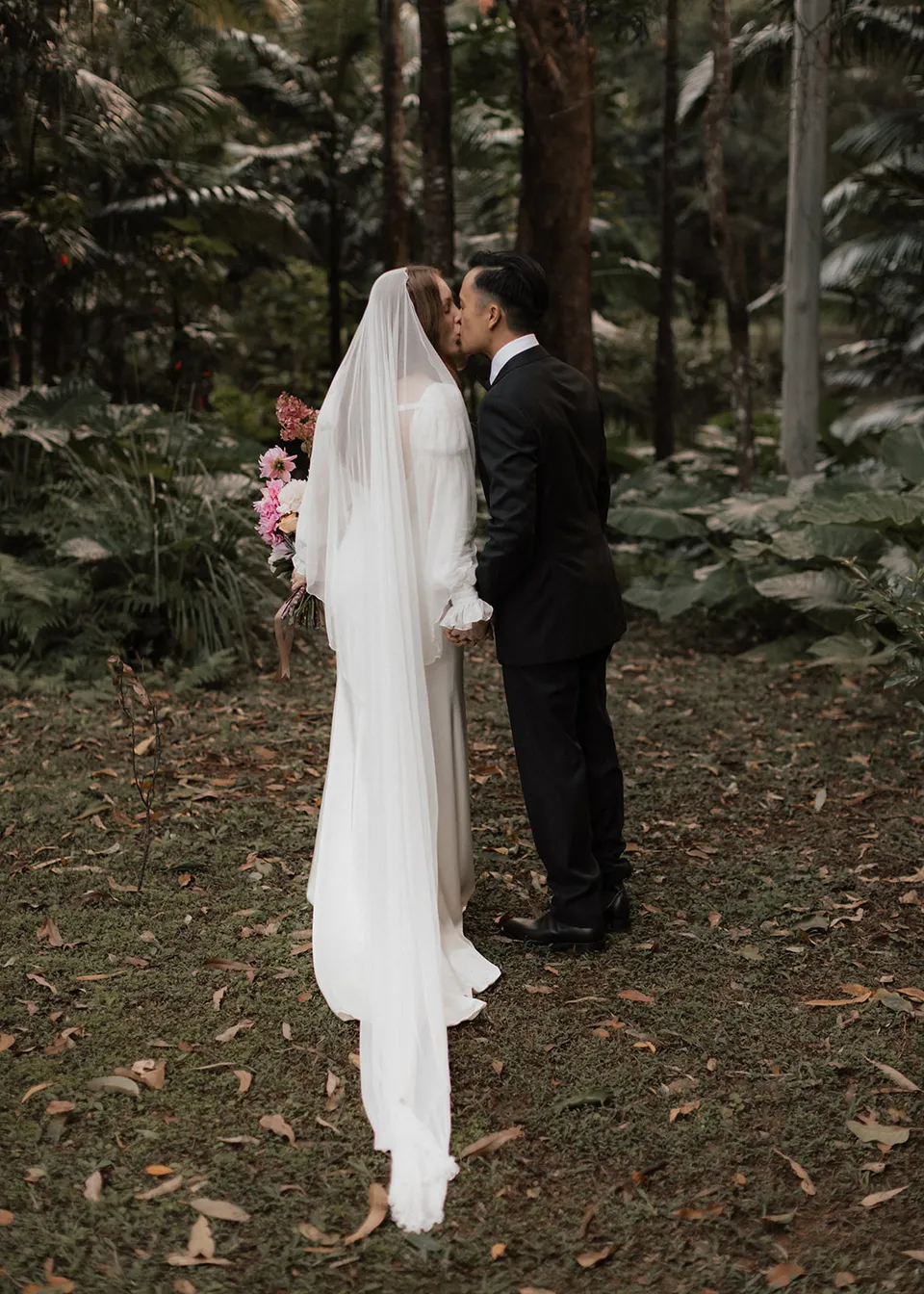 Jonathan kissing Elyse's cheek among the tall trees. At Fig Tree Villa in Ewingsdale, Byron Bay, for an Elopement Collective elopement. Photograph by Bulb Creative.