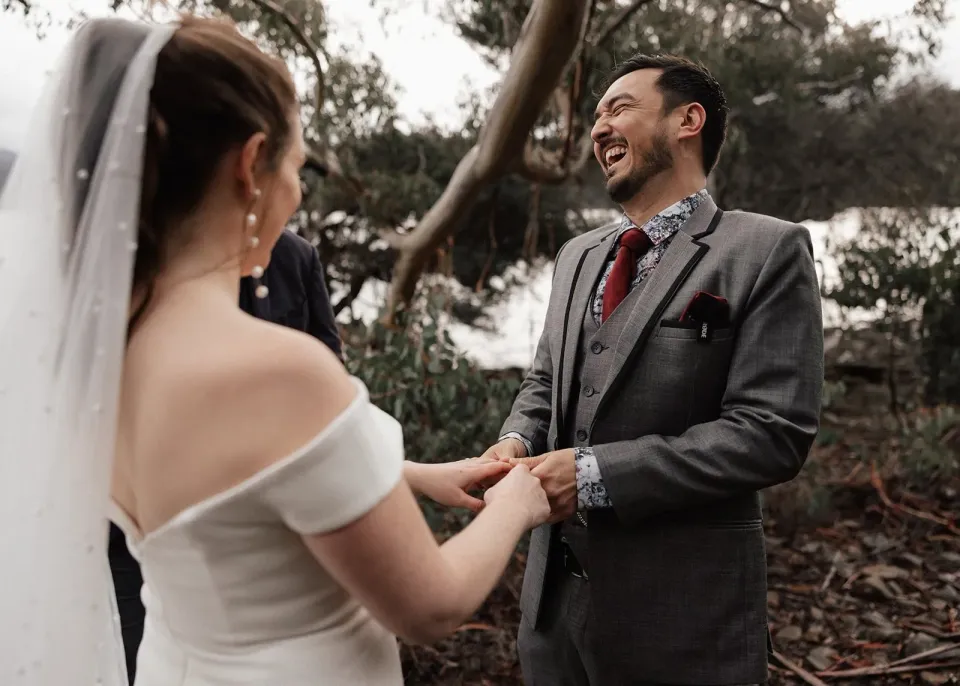 Jono laughing as he and Sarah hold hands beneath the lakeside trees, at Pumphouse Point, Lake St Clair, Tasmania, for an Elopement Collective elopement. Photo by Bulb Creative.