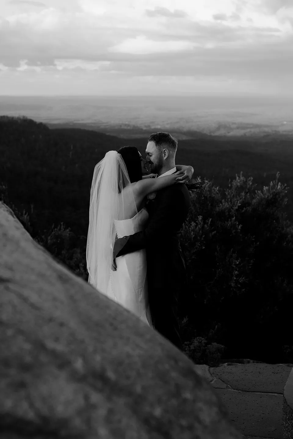 Black and white photograph of Meg and Ryan embracing on the rocky hilltop above layers of distant hills. At The Keep on Tasmania’s east coast, for an Elopement Collective elopement. Photograph by Jenna Agius.