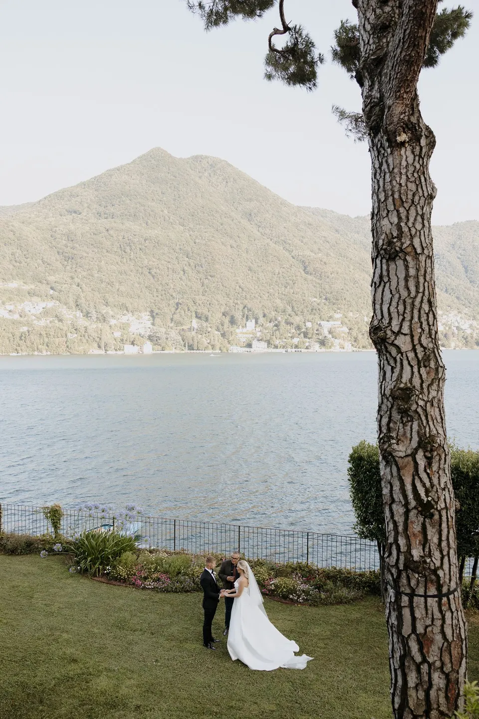 Wide view of Zoe and Charlton on the lakeside ceremony lawn, framed by the trunk of a tall tree and the wooded mountains across the water, at Passalacqua in Moltrasio, Lake Como, Italy, for an Elopement Collective elopement. Photo by George Bowden.