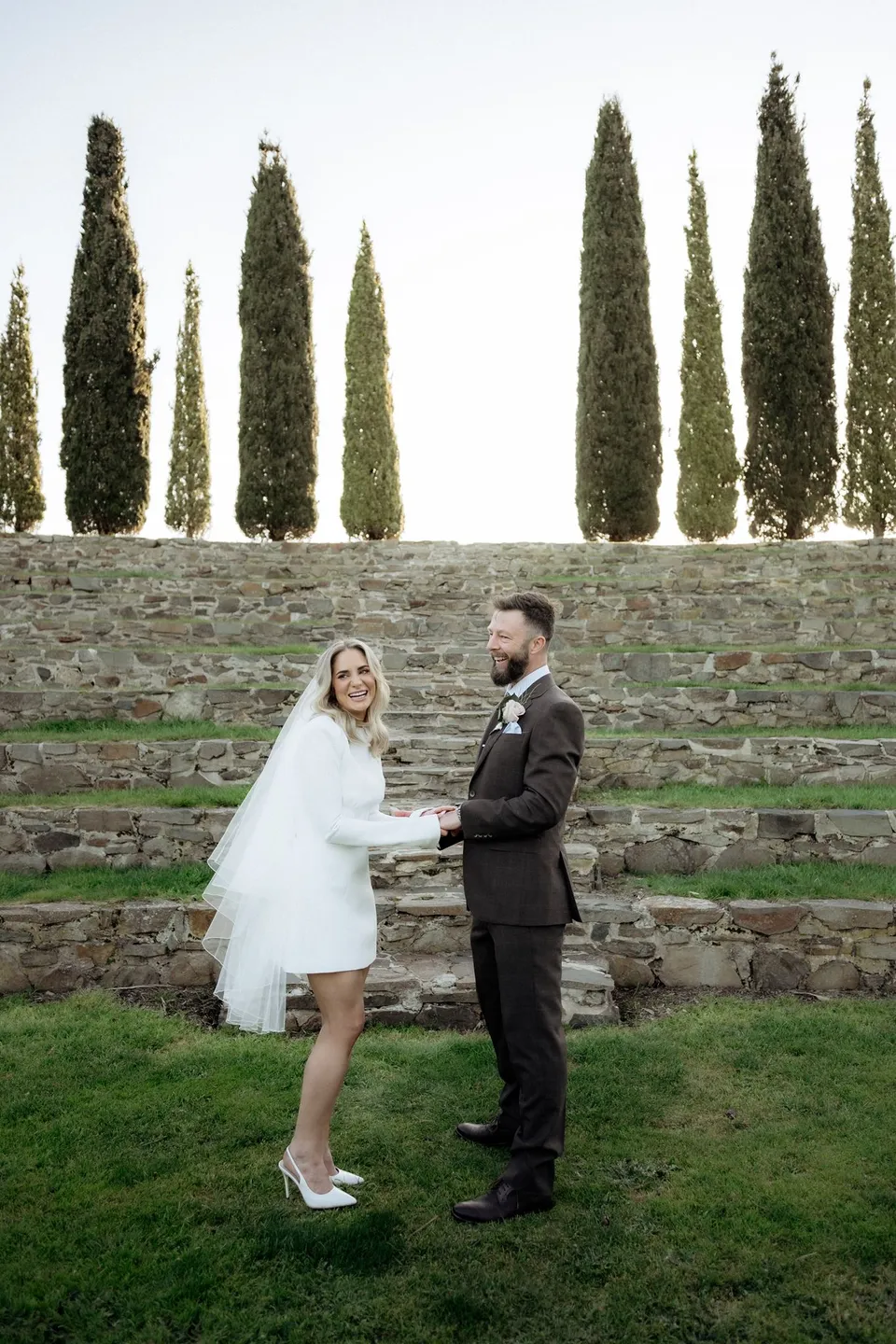 Kaitlyn and Russell holding hands and smiling on the grass in front of the stone amphitheatre. At Piermont Retreat on Tasmania’s east coast, for an Elopement Collective elopement. Photograph by Omar of By The Wilde.