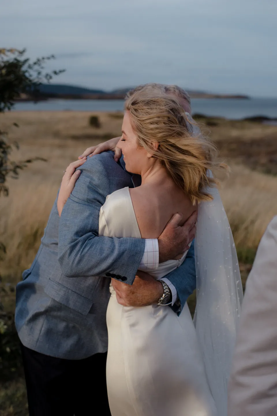 Jemmah embracing a loved one with the meadow behind them, at Thalia Haven on the East Coast of Tasmania, Australia, for an Elopement Collective elopement. Photograph by Joey Willis.