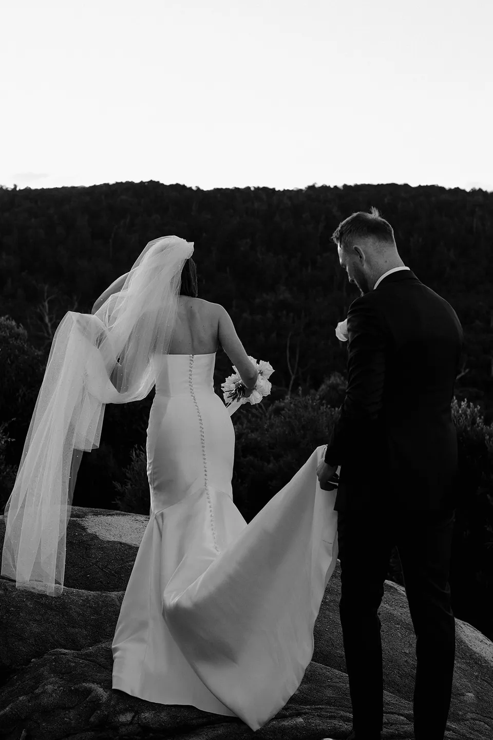 Black and white photograph of Ryan helping carry Meg’s wedding dress train as they walk across the rocks. At The Keep on Tasmania’s east coast, for an Elopement Collective elopement. Photograph by Jenna Agius.