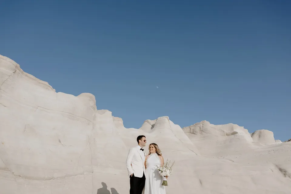 Georgina and Brad standing together against a sculpted white rock wall and blue sky, with her white Giasemi bouquet, at Sarakiniko on Milos in the Cyclades, Greece, during an Elopement Collective elopement. Photograph by George Bowden.
