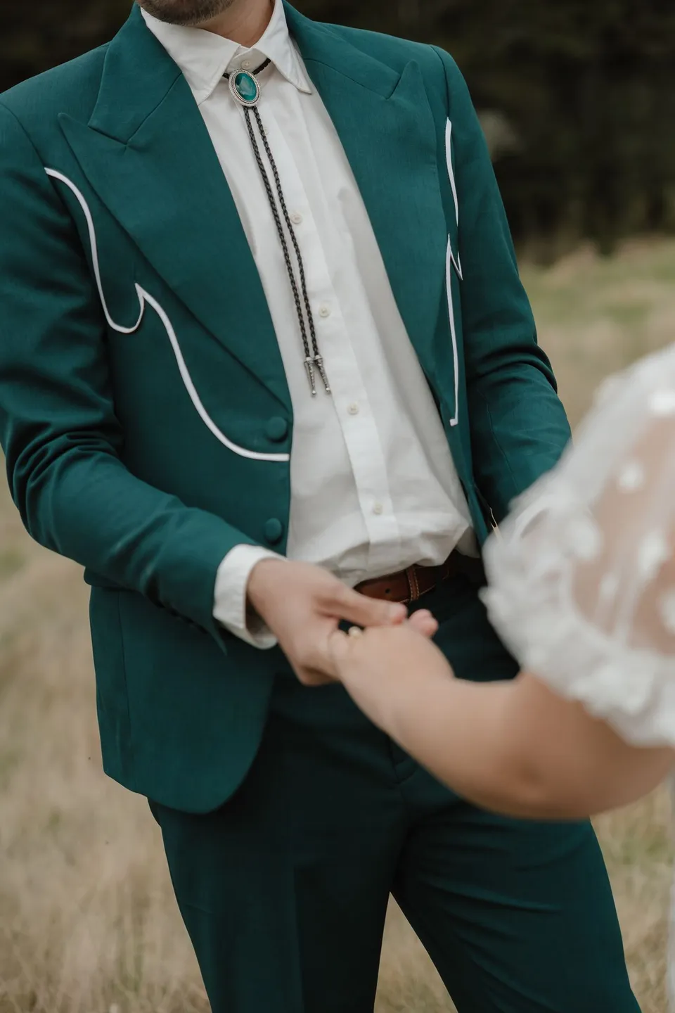A close view of Jaeda and Adrian holding hands during their ceremony, with his green suit and turquoise bolo tie visible, in the field on the road to Paradise near Glenorchy, in the Queenstown region of New Zealand, for an Elopement Collective Queenstown elopement. Photograph by Bulb Creative.