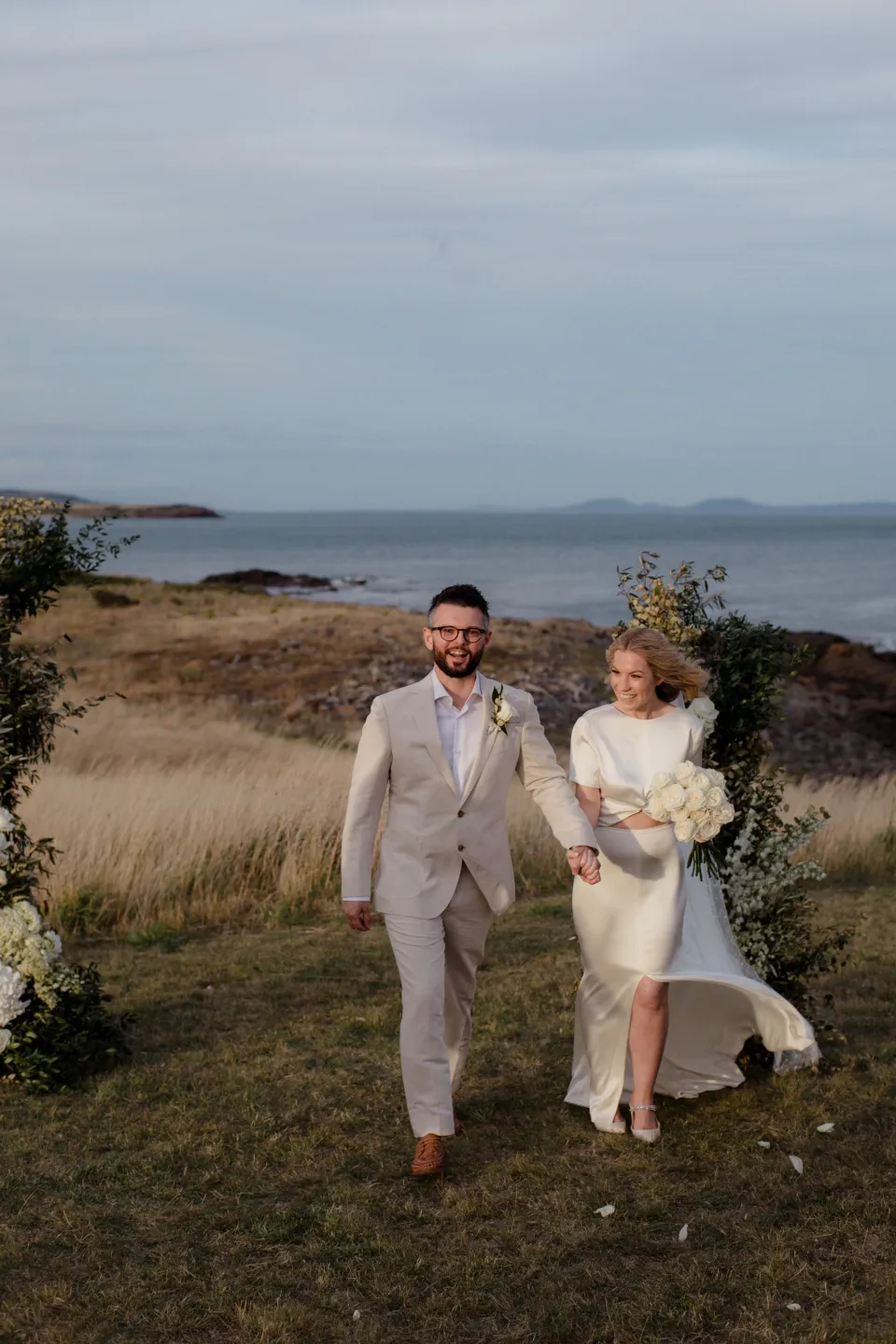 Jemmah and Jack walking hand in hand from their ceremony, at Thalia Haven on the East Coast of Tasmania, Australia, for an Elopement Collective elopement. Photograph by Joey Willis.