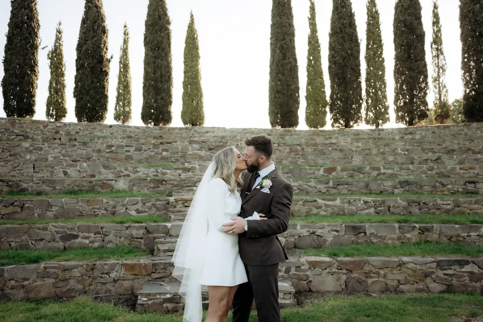 Kaitlyn and Russell kissing beneath the cypress trees, with curved stone amphitheatre seating behind them. At Piermont Retreat on Tasmania’s east coast, for an Elopement Collective elopement. Photograph by Omar of By The Wilde.
