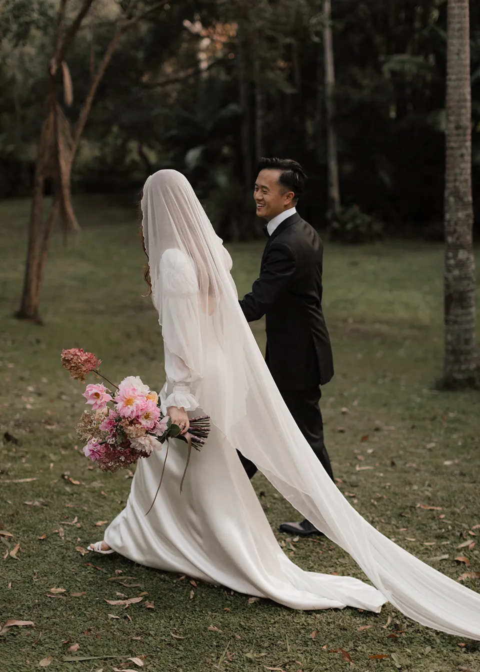 Elyse and Jonathan looking back towards the camera beneath the trees. At Fig Tree Villa in Ewingsdale, Byron Bay, for an Elopement Collective elopement. Photograph by Bulb Creative.