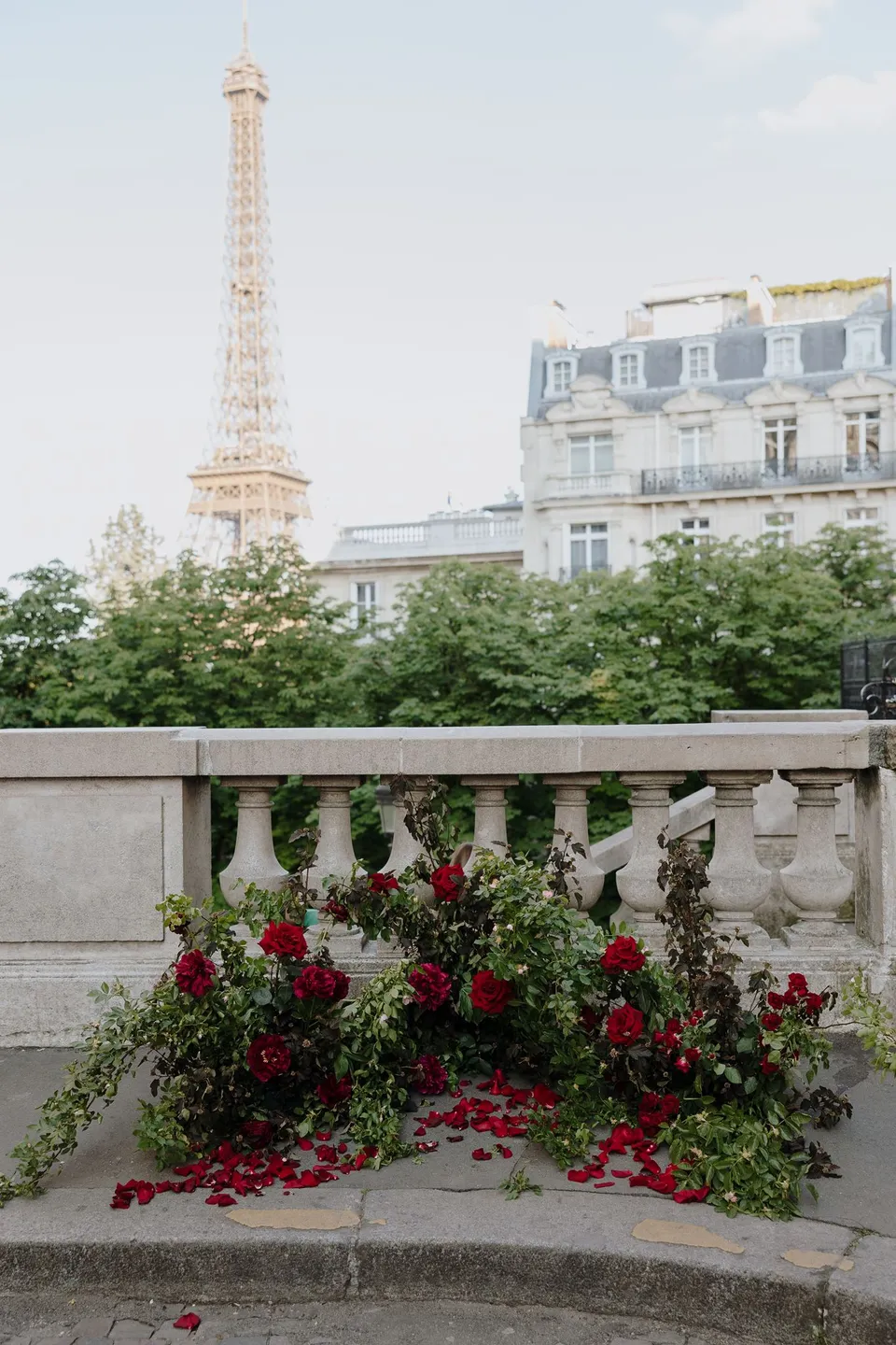 Ivy Bleu’s red floral ceremony arrangements beside a stone balustrade, with the Eiffel Tower beyond the trees, at Avenue de Camoëns in Paris, France, during an Elopement Collective elopement. Photograph by George Bowden.