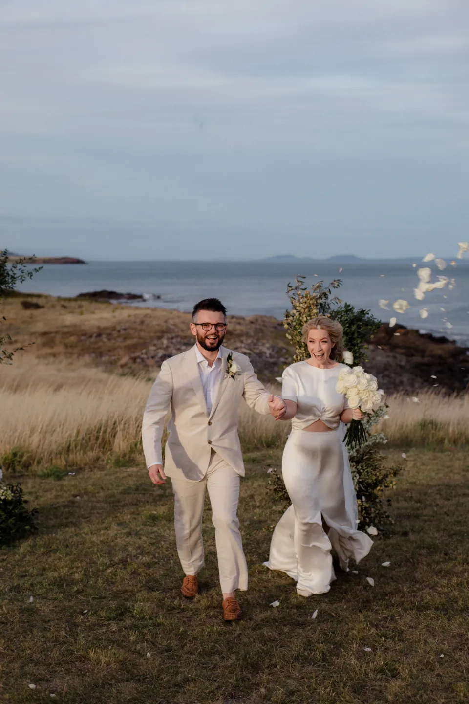 The newlyweds walking towards the camera with Jemmah’s bouquet by The Romantics, at Thalia Haven on the East Coast of Tasmania, Australia, for an Elopement Collective elopement. Photograph by Joey Willis.