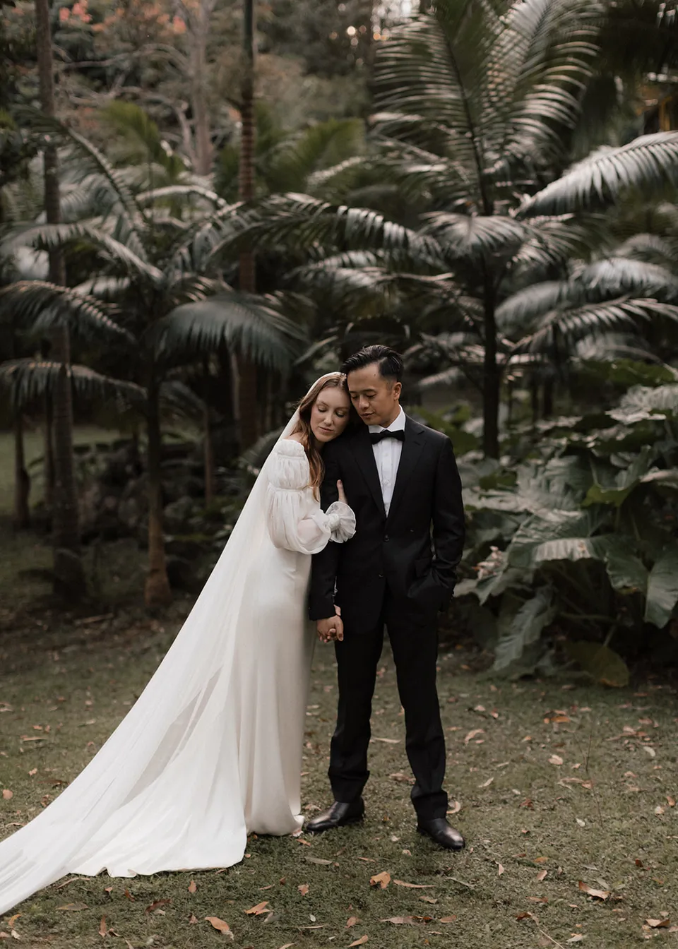 Elyse leaning against Jonathan with her long veil spread across the grass. At Fig Tree Villa in Ewingsdale, Byron Bay, for an Elopement Collective elopement. Photograph by Bulb Creative.