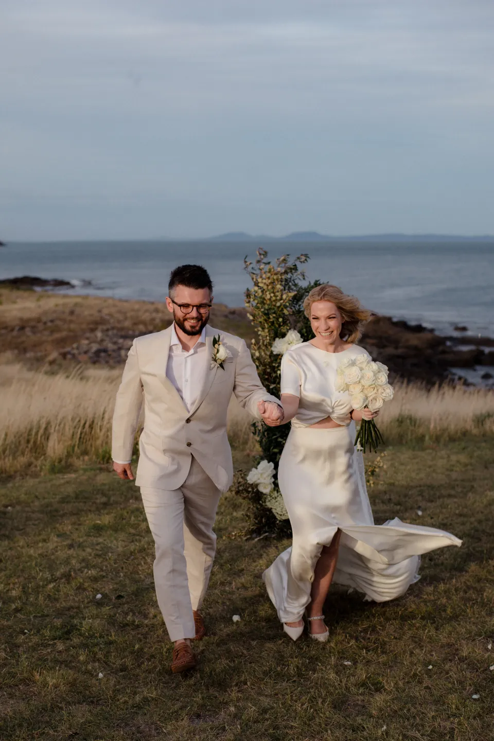 Jemmah and Jack walking through the meadow with her dress moving in the wind, at Thalia Haven on the East Coast of Tasmania, Australia, for an Elopement Collective elopement. Photograph by Joey Willis.