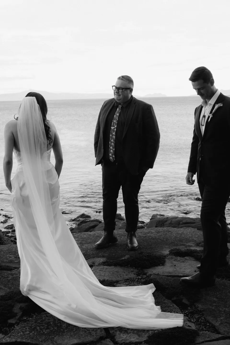 Black and white photograph of Dani and Anthony with celebrant Josh Withers on coastal rocks beside the bay, at Thalia Haven on Tasmania’s east coast, for an Elopement Collective elopement. Photograph by Bulb Creative.
