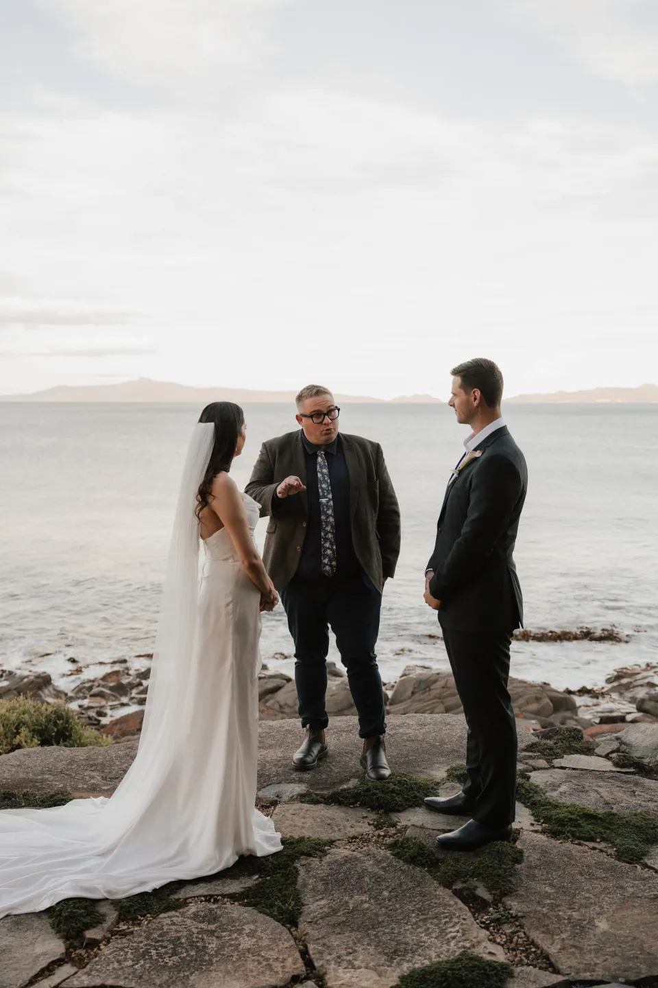 Dani and Anthony standing with celebrant Josh Withers for their intimate ceremony on the rocky shoreline, at Thalia Haven on Tasmania’s east coast, for an Elopement Collective elopement. Photograph by Bulb Creative.