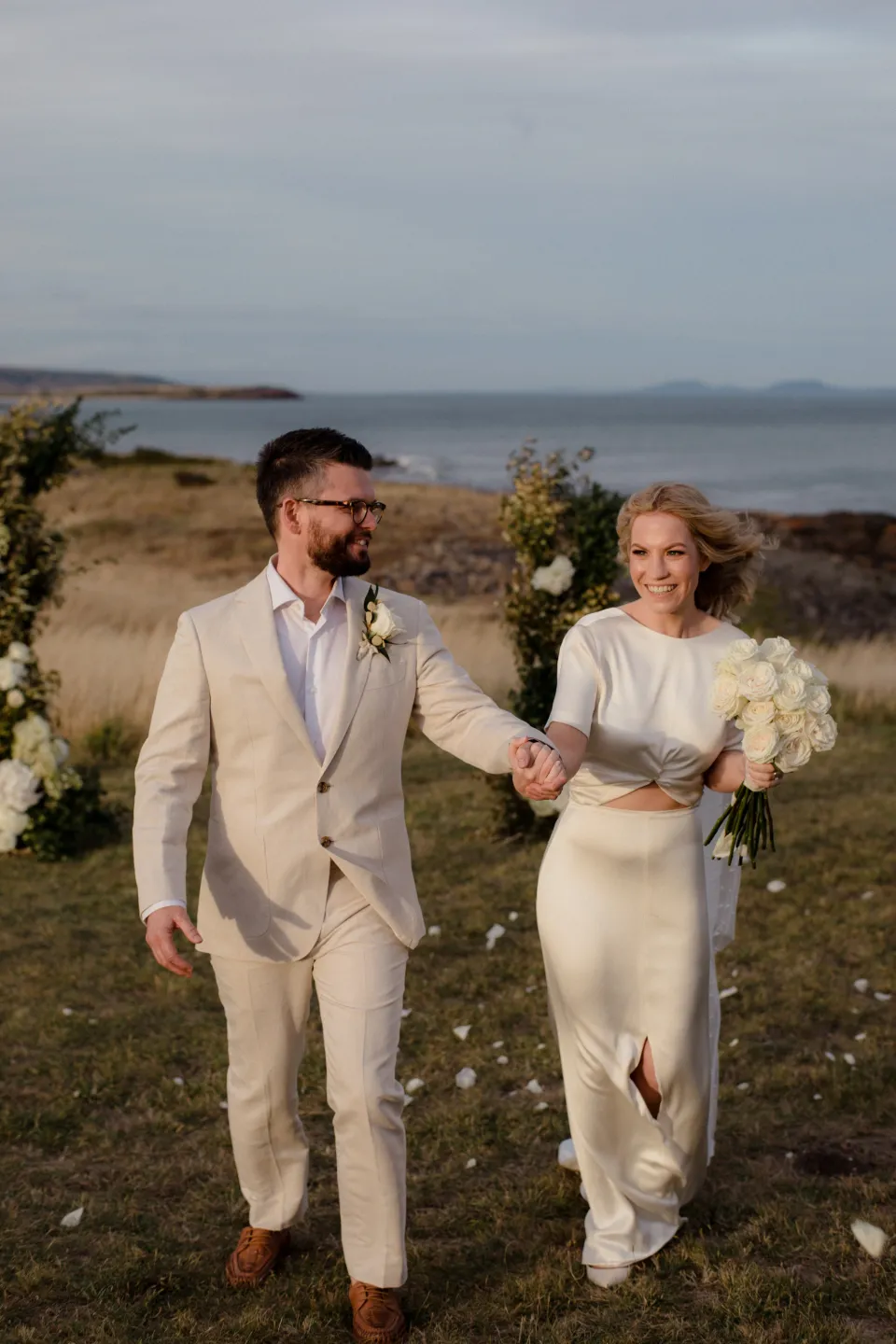 The couple walking hand in hand with the bay behind them, at Thalia Haven on the East Coast of Tasmania, Australia, for an Elopement Collective elopement. Photograph by Joey Willis.