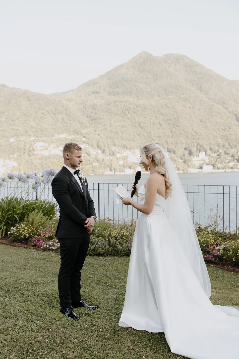 Zoe reading her vows into a microphone as Charlton listens, with the lake and mountain villages behind them, at Passalacqua in Moltrasio, Lake Como, Italy, for an Elopement Collective elopement. Photo by George Bowden.