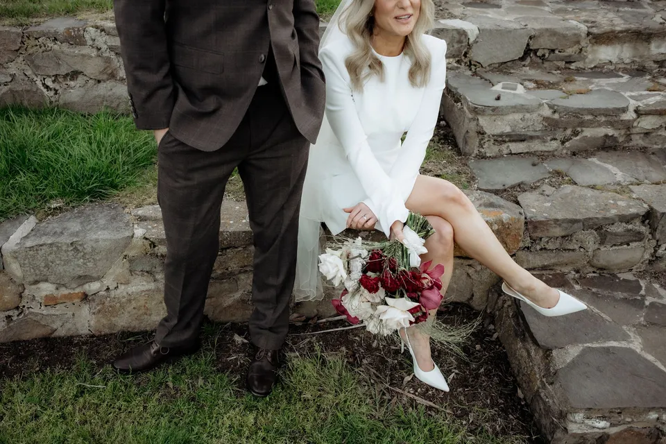 Kaitlyn’s cream and burgundy bouquet by The Romantics beside her white shoes as she sits on the amphitheatre steps. At Piermont Retreat on Tasmania’s east coast, for an Elopement Collective elopement. Photograph by Omar of By The Wilde.