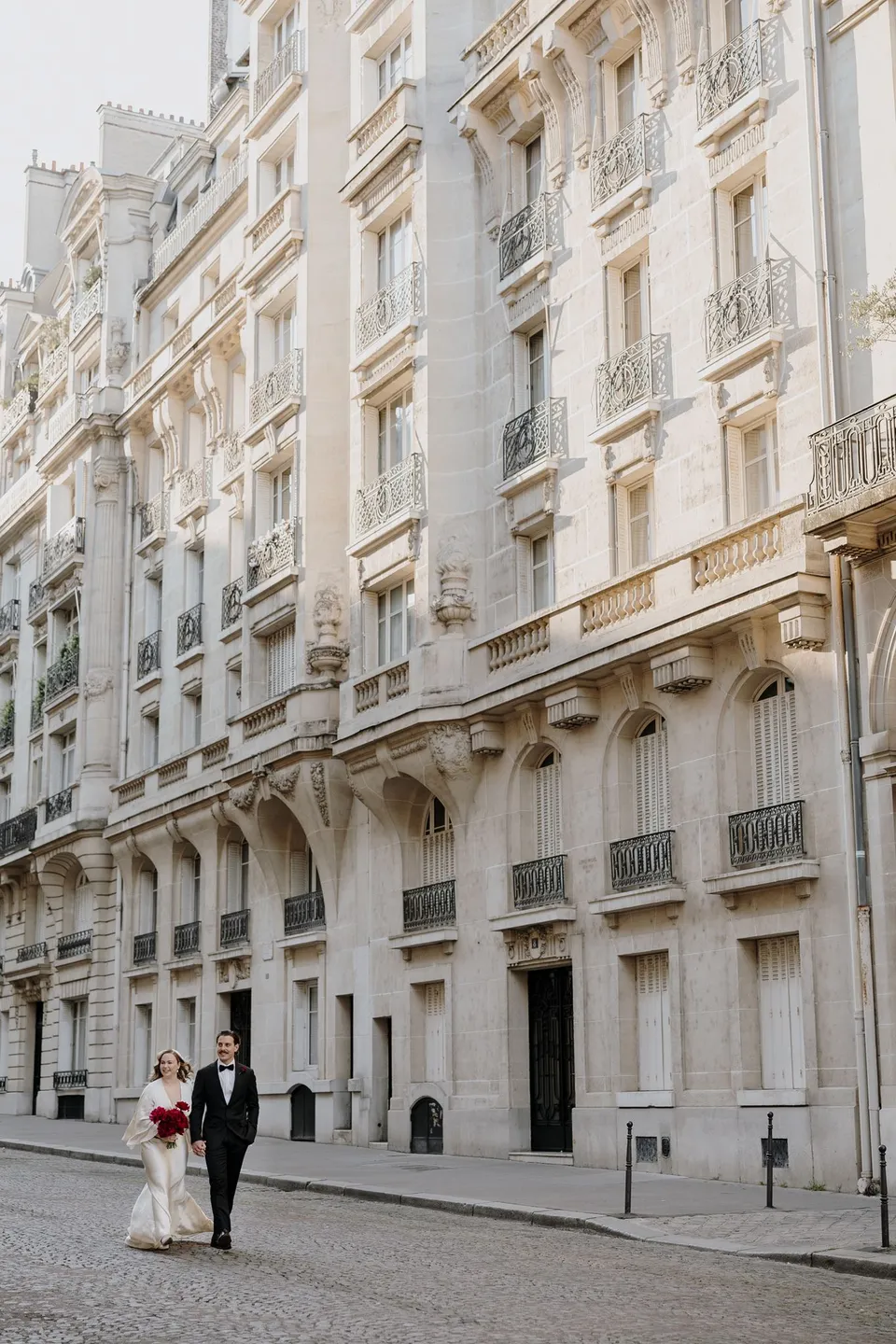 Kara and Samuel walking together along a cobbled street beside ornate cream facades, with her red Ivy Bleu bouquet, at Avenue de Camoëns in Paris, France, during an Elopement Collective elopement. Photograph by George Bowden.
