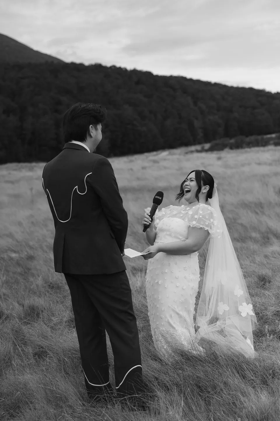 A black and white photograph of Jaeda and Adrian facing each other and holding hands in the meadow, in the field on the road to Paradise near Glenorchy, in the Queenstown region of New Zealand, for an Elopement Collective Queenstown elopement. Photograph by Bulb Creative.