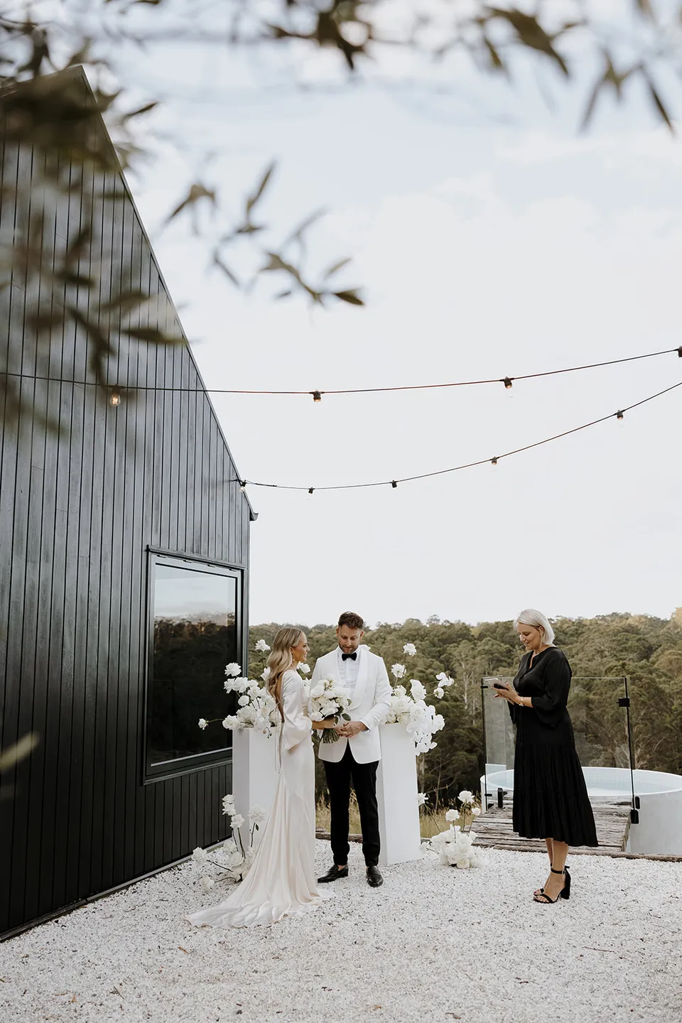 Wide view of Gabrielle and Greg with celebrant Hayley Hart beside the cabin, white flowers by Mrs Gibbons and hinterland trees, at Roundhill Retreat in Obi Obi, Sunshine Coast Hinterland, Queensland, for an Elopement Collective elopement. Photo by George Bowden.