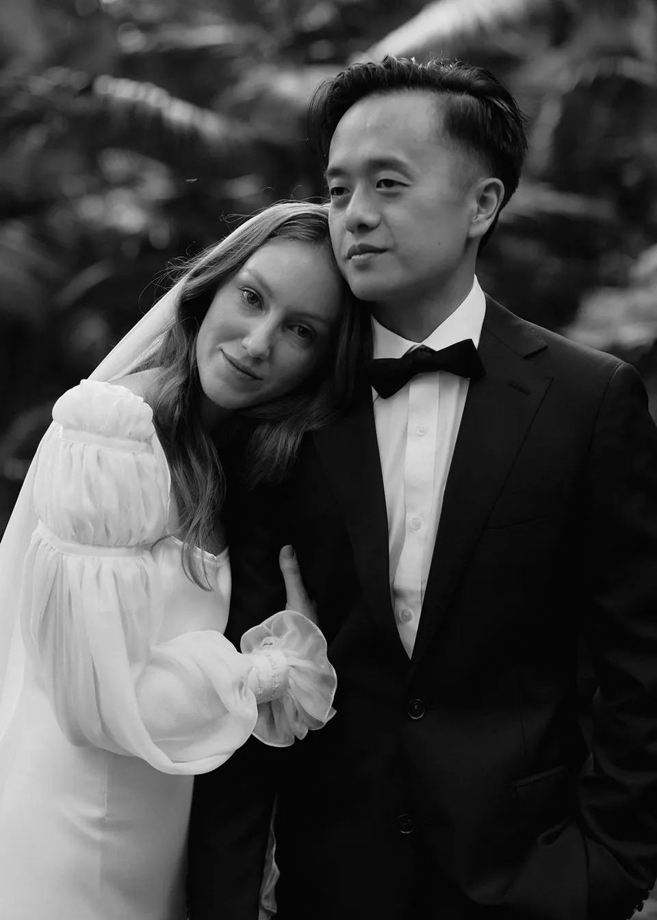 Black-and-white portrait of Elyse leaning against Jonathan in the garden. At Fig Tree Villa in Ewingsdale, Byron Bay, for an Elopement Collective elopement. Photograph by Bulb Creative.