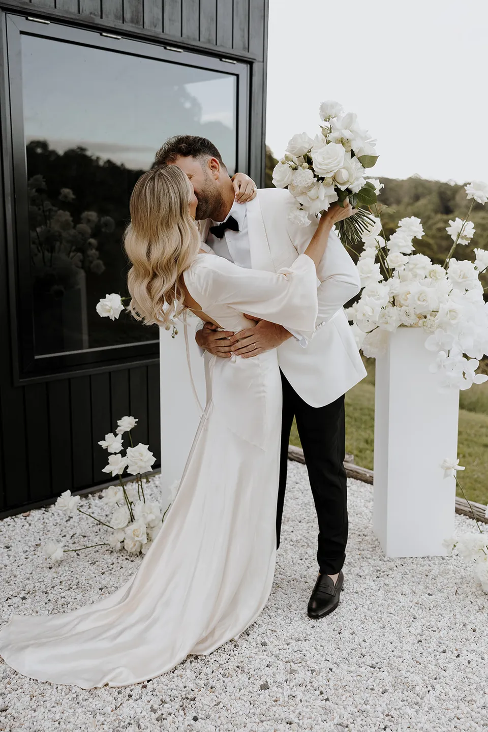 Greg embracing Gabrielle as they kiss beside the ceremony flowers, her white bouquet by Mrs Gibbons raised in her hand, at Roundhill Retreat in Obi Obi, Sunshine Coast Hinterland, Queensland, for an Elopement Collective elopement. Photo by George Bowden.