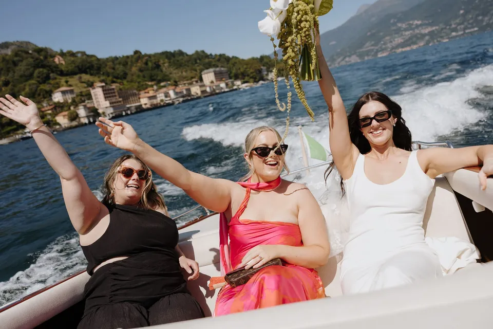 Lyndal and two guests smiling and raising their hands aboard a boat, with her white bouquet by Garden of Love and the shoreline behind them, at Lake Como in Lombardy, Italy, for an Elopement Collective elopement. Photograph by George Bowden.