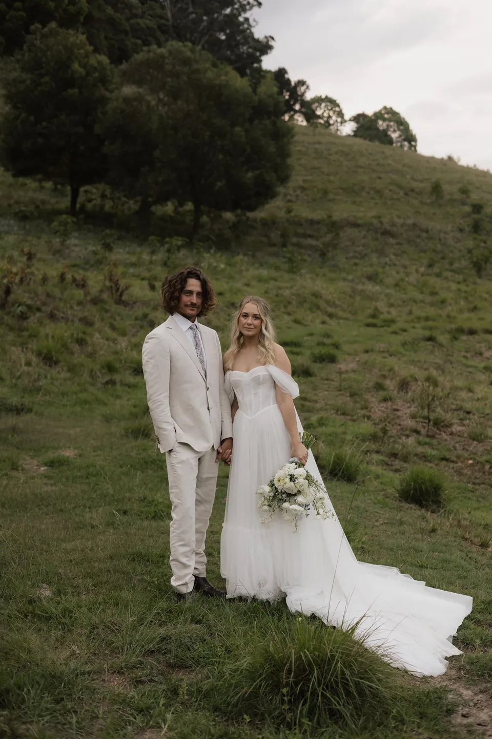 Bridget and James standing together in the grass, her bouquet and long train beside them. At Cudgera Creek in northern New South Wales, for an Elopement Collective elopement. Photograph by Bulb Creative.