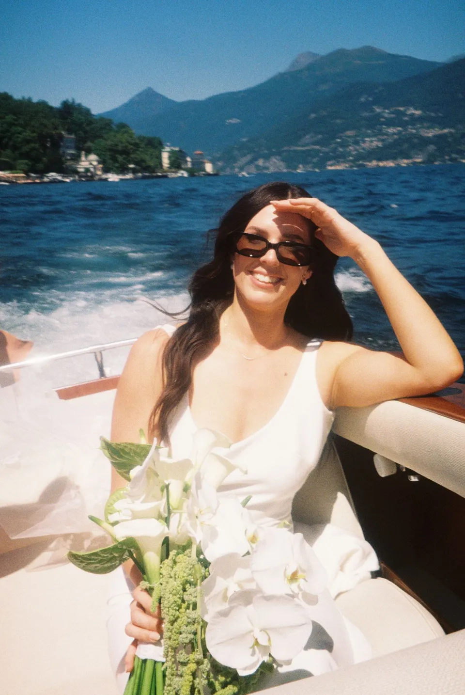 Lyndal seated on a boat in her white halter dress and sunglasses, with a bouquet by Garden of Love on her lap and the wake behind her, at Lake Como in Lombardy, Italy, for an Elopement Collective elopement. Photograph by George Bowden.